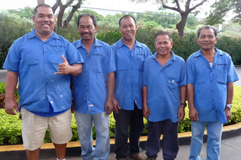 Five men in blue shirts posing outdoors with greenery in the background.