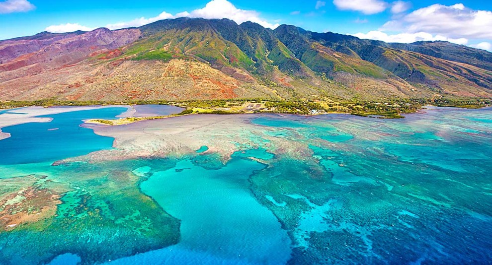 Aerial view of turquoise waters with coral reefs and mountains in the background.